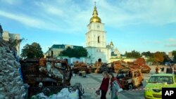 Two women holding bouquets of flowers stand in front of a display of destroyed Russian tanks and armored vehicles near St. Michael's Golden-Domed Monastery in Kyiv, Sept. 11, 2022