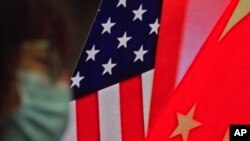 Seorang perempuan mengenakan masker duduk dekat layar televisi yang menampilkan bendera AS dan China untuk mendengarkan pidato mengenai hubungan AS-China, 22 Februari 2022. (Foto: Andy Wong/AP)