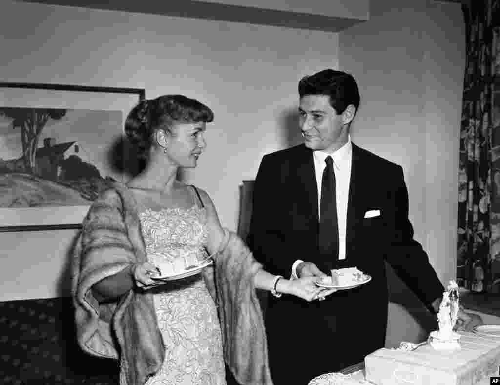 តារាភាពយន្ត Debbie Reynolds ហុចនំខេកទៅឲ្យស្វាមីរបស់អ្នកស្រី លោក Eddie Fisher ដែលជាអ្នកចម្រៀង នៅពេលដែលពួកគេឈរថតរូបនៅក្នុងសន្និសីទកាសែតនៅរដ្ឋធានី Washington, D.C., ប្រហែលជា២៤ម៉ោង ក្រោយពីពួកគេរៀបអាពាហ៍ពិពាហ៍រួចនៅរមណីយដ្ឋានមួយនៅភ្នំ Catskill កាលពីថ្ងៃទី២៧ កញ្ញា ១៩៥៥។