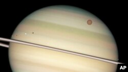 FILE - This image provided by NASA captures the transit of several moons across the face of Saturn. The giant orange moon Titan – larger than the planet Mercury – can be seen at upper right.