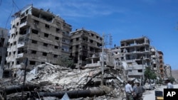 Warga berdiri di depan gedung yang hancur di Douma, dekat Damaskus, Suriah, pada 16 April 2018. Gedung tersebut diduga menjadi bagian dari lokasi serang senjata kimia dalam Perang Suriah. (Foto: AP/Hassan Ammar)