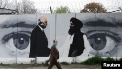 FILE - An Afghan man wearing a protective face mask walks past a wall painted with images of Zalmay Khalilzad, U.S. envoy in Afghanistan, and Mullah Abdul Ghani Baradar, the leader of the Taliban delegation, in Kabul, Afghanistan, April 13, 2020.